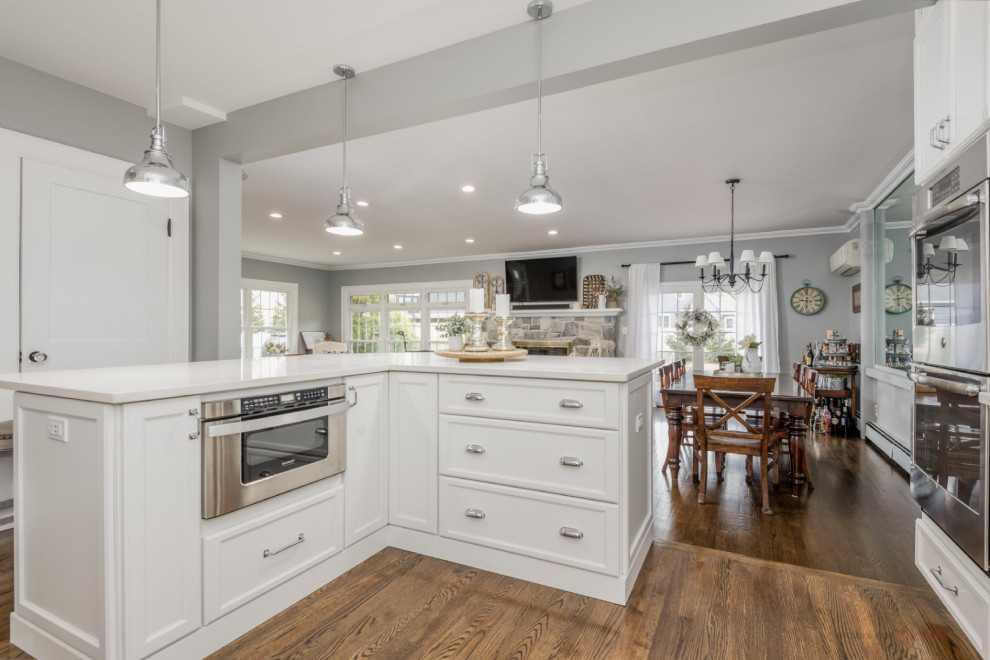 Photo of a mid-sized traditional l-shaped eat-in kitchen in New York with a farmhouse sink, recessed-panel cabinets, white cabinets, quartz benchtops, grey splashback, stainless steel appliances, dark hardwood floors, with island, brown floor and white benchtop.
