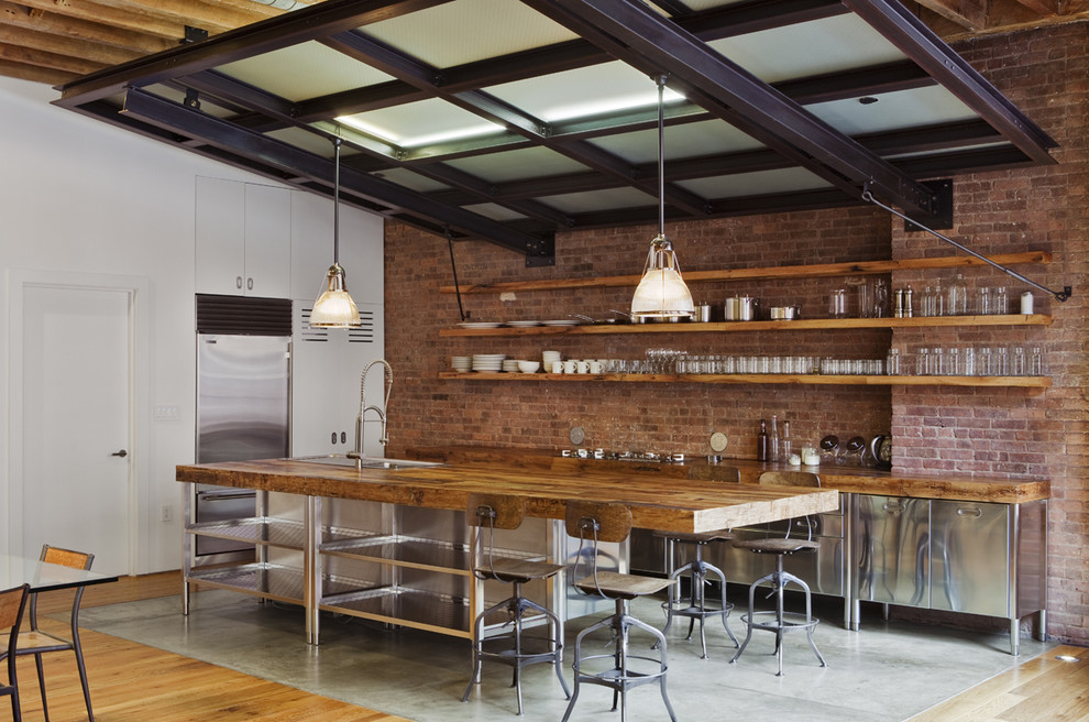 Inspiration for a mid-sized industrial l-shaped eat-in kitchen in New York with stainless steel appliances, stainless steel cabinets, wood benchtops, flat-panel cabinets, red splashback, brick splashback, with island, a farmhouse sink, concrete floors and grey floor.