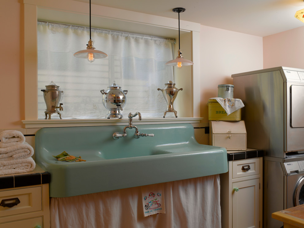 Design ideas for a mid-sized traditional galley dedicated laundry room in Seattle with a farmhouse sink, recessed-panel cabinets, white cabinets, tile benchtops, pink walls, light hardwood floors and a stacked washer and dryer.