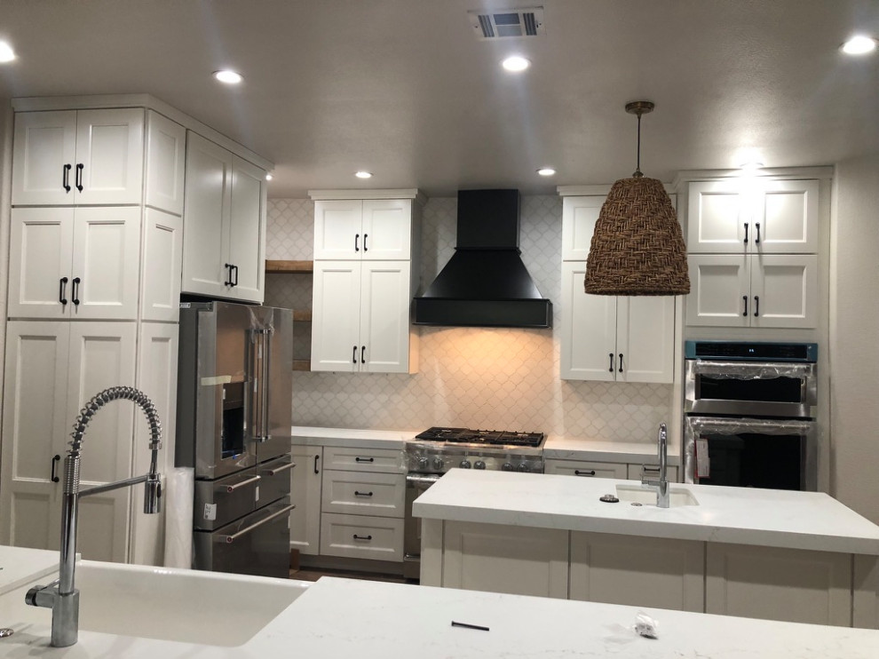 This is an example of a large contemporary l-shaped eat-in kitchen in Las Vegas with a farmhouse sink, shaker cabinets, white cabinets, limestone benchtops, white splashback, marble splashback, stainless steel appliances, light hardwood floors, multiple islands, brown floor and white benchtop.