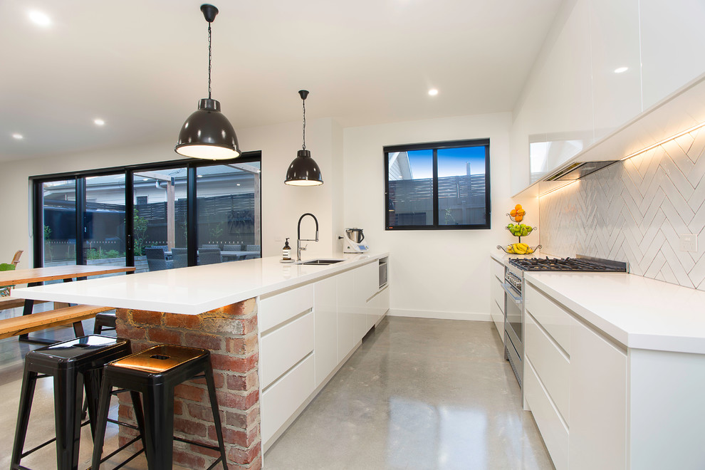 This is an example of a mid-sized contemporary single-wall open plan kitchen in Melbourne with an undermount sink, flat-panel cabinets, white cabinets, solid surface benchtops, white splashback, stainless steel appliances, concrete floors, a peninsula and grey floor.