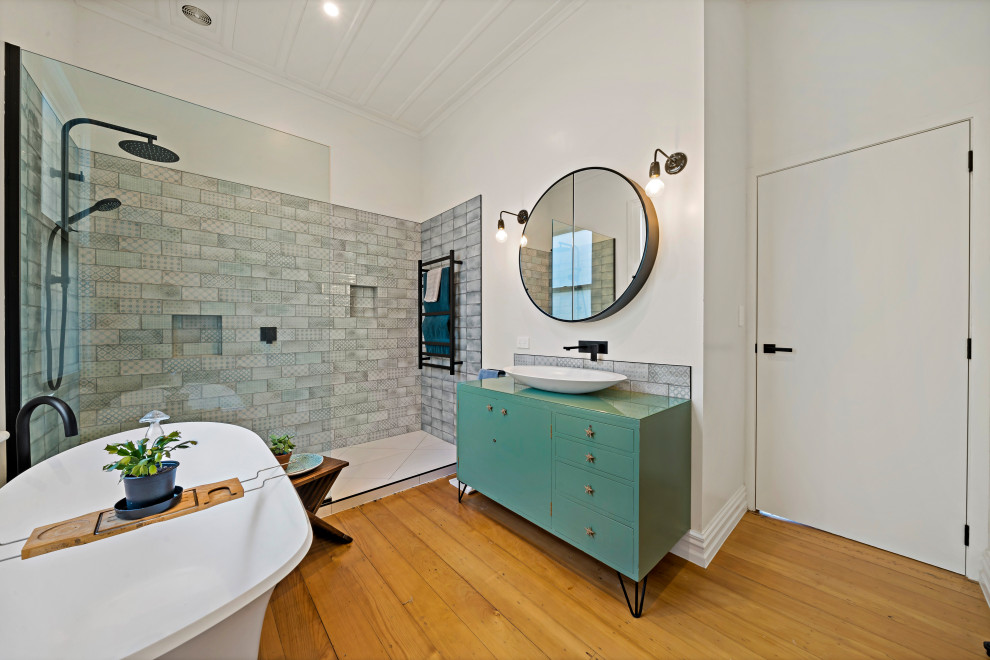 Inspiration for a large eclectic master bathroom in Auckland with furniture-like cabinets, green cabinets, a freestanding tub, an open shower, a wall-mount toilet, gray tile, subway tile, white walls, medium hardwood floors, a vessel sink, glass benchtops, brown floor, an open shower, green benchtops, a single vanity, a freestanding vanity and exposed beam.
