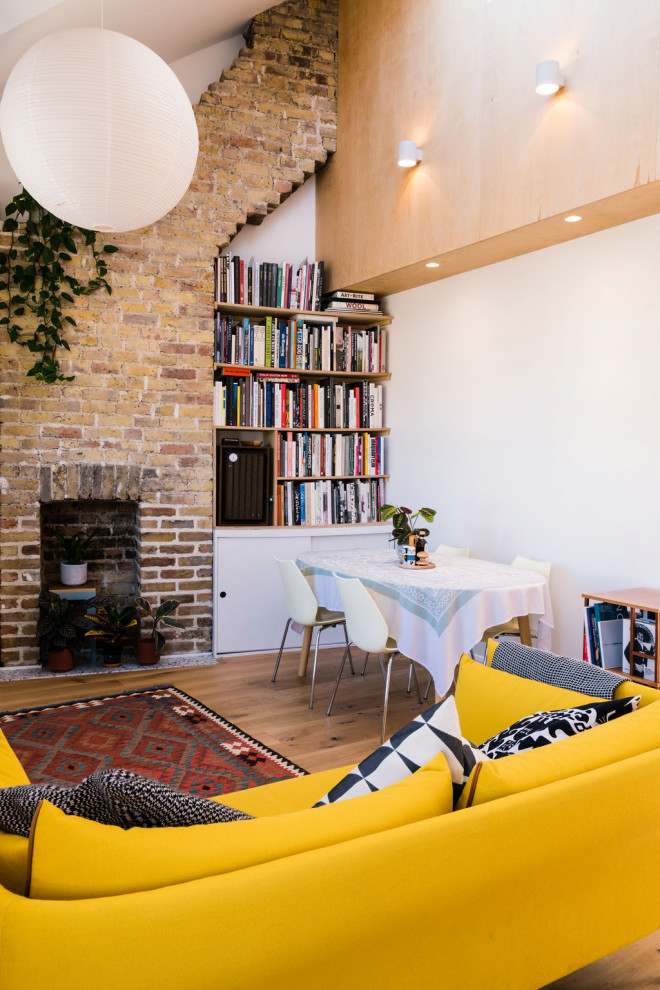 This is an example of a mid-sized contemporary loft-style living room in London with white walls, light hardwood floors, a brick fireplace surround, vaulted and brick walls.