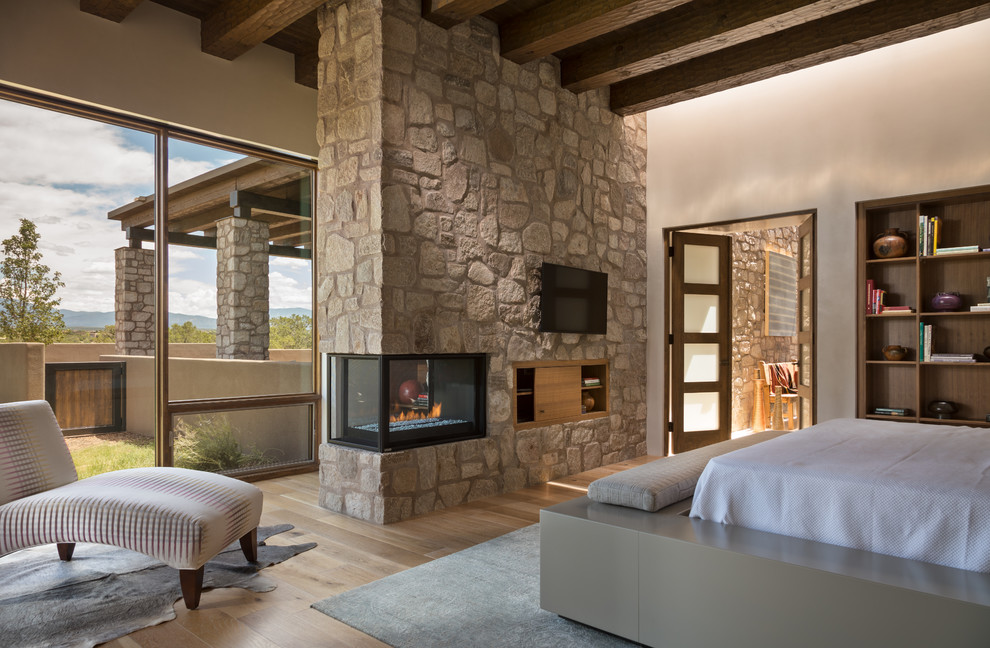 This is an example of a large master bedroom in Albuquerque with beige walls, a corner fireplace, a stone fireplace surround, brown floor and light hardwood floors.