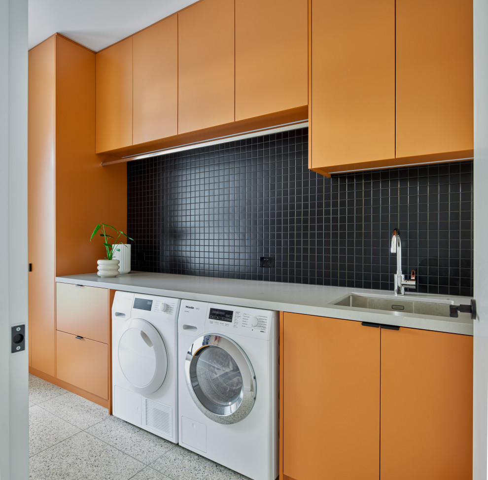 This is an example of a large contemporary single-wall dedicated laundry room in Melbourne with an undermount sink, flat-panel cabinets, orange cabinets, solid surface benchtops, porcelain floors, a side-by-side washer and dryer, white walls and grey benchtop.