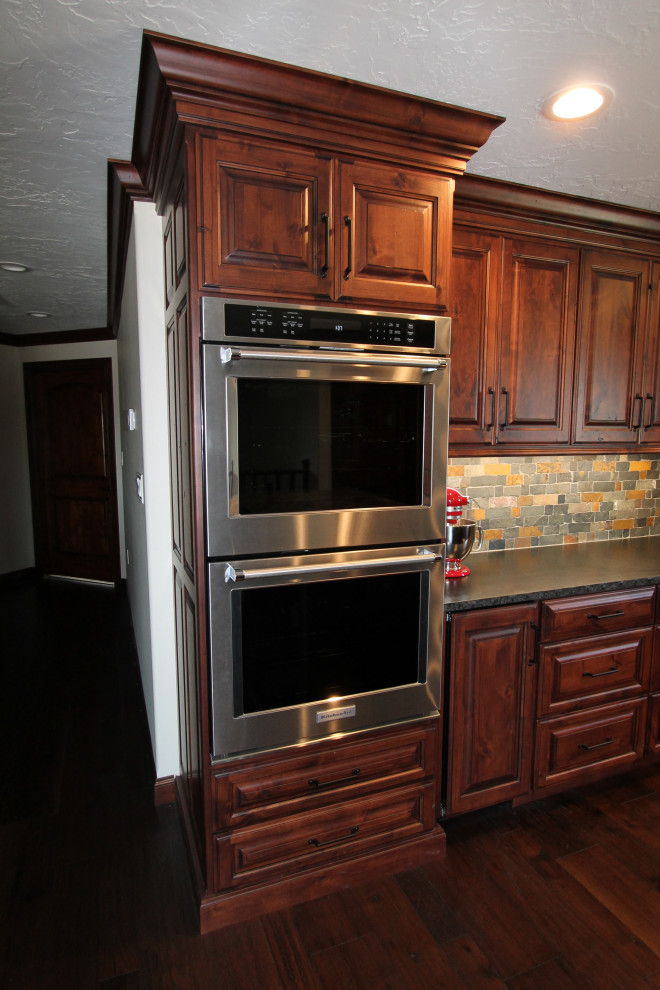 Kitchen - large rustic medium tone wood floor and brown floor kitchen idea with raised-panel cabinets, medium tone wood cabinets, granite countertops, multicolored backsplash, stone tile backsplash, paneled appliances, an island and black countertops