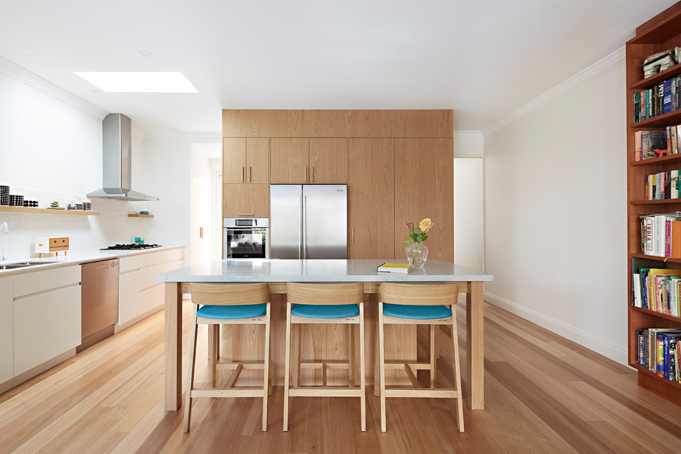Inspiration for a large contemporary single-wall open plan kitchen in Melbourne with a double-bowl sink, white splashback, subway tile splashback, stainless steel appliances, light hardwood floors, with island, beaded inset cabinets, medium wood cabinets, marble benchtops, brown floor and white benchtop.