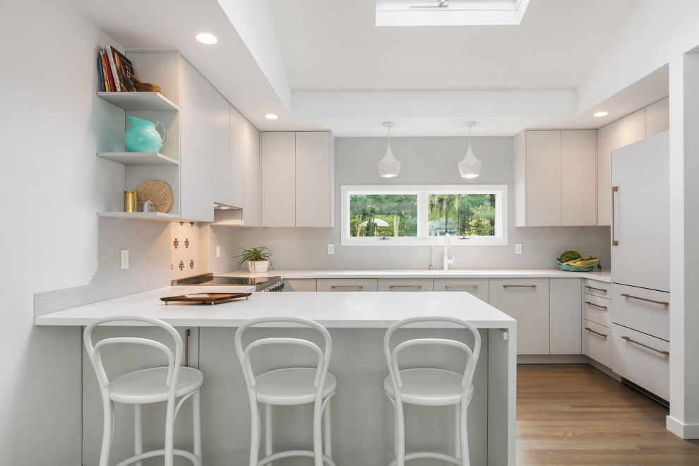 This is an example of a beach style u-shaped kitchen in New York with flat-panel cabinets, white cabinets, panelled appliances, medium hardwood floors, a peninsula, brown floor and white benchtop.