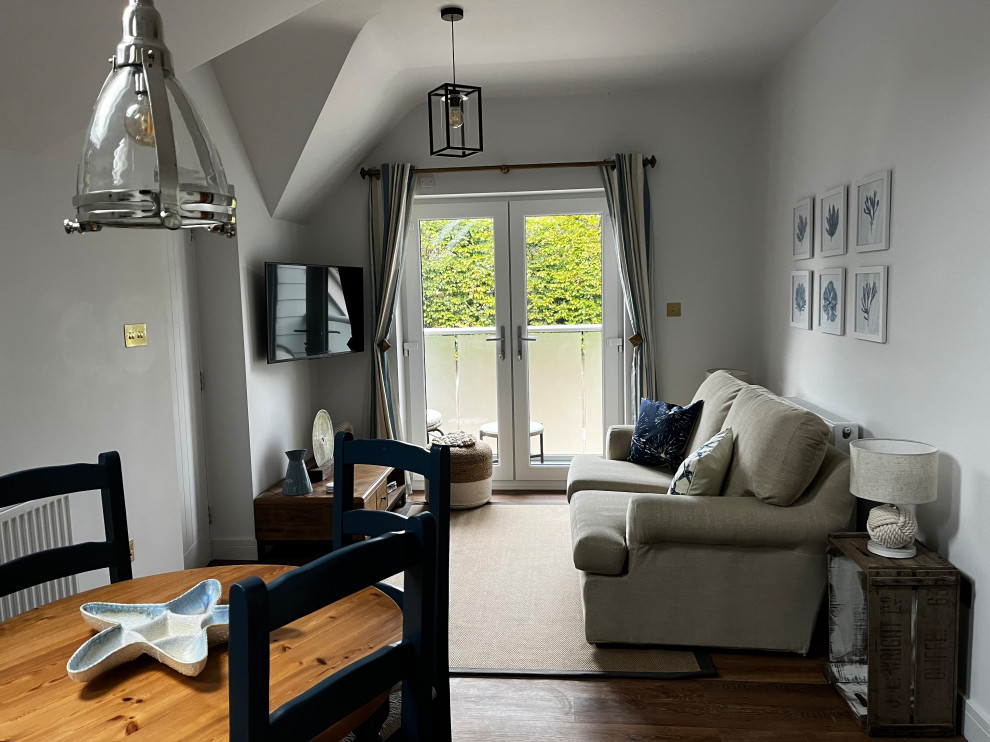 На фото: открытая гостиная комната среднего размера:: освещение в морском стиле с белыми стенами, полом из ламината, телевизором на стене, коричневым полом и сводчатым потолком