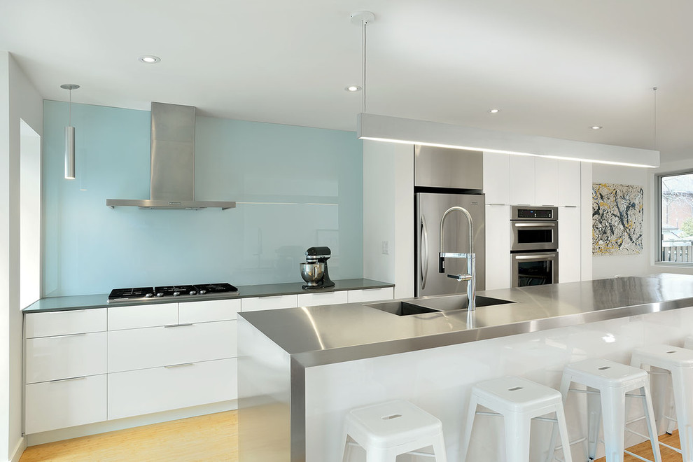 Photo of a contemporary galley kitchen in Toronto with an integrated sink, flat-panel cabinets, white cabinets, stainless steel benchtops, blue splashback, glass sheet splashback and stainless steel appliances.