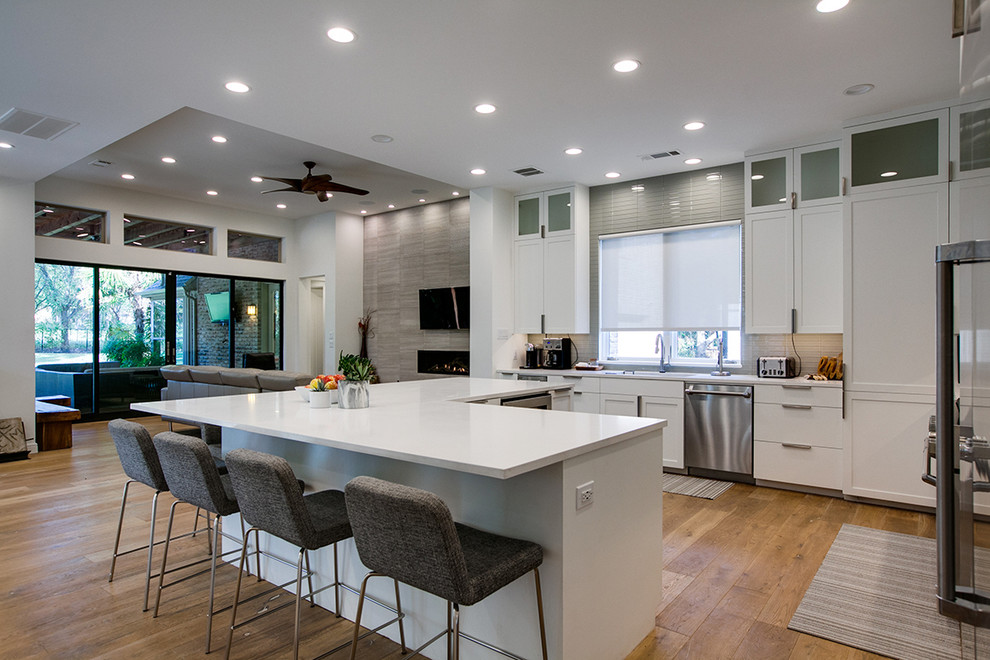 This is an example of a large contemporary l-shaped open plan kitchen in Dallas with shaker cabinets, white cabinets, stainless steel appliances, with island, an undermount sink, quartzite benchtops, beige splashback, glass tile splashback and light hardwood floors.