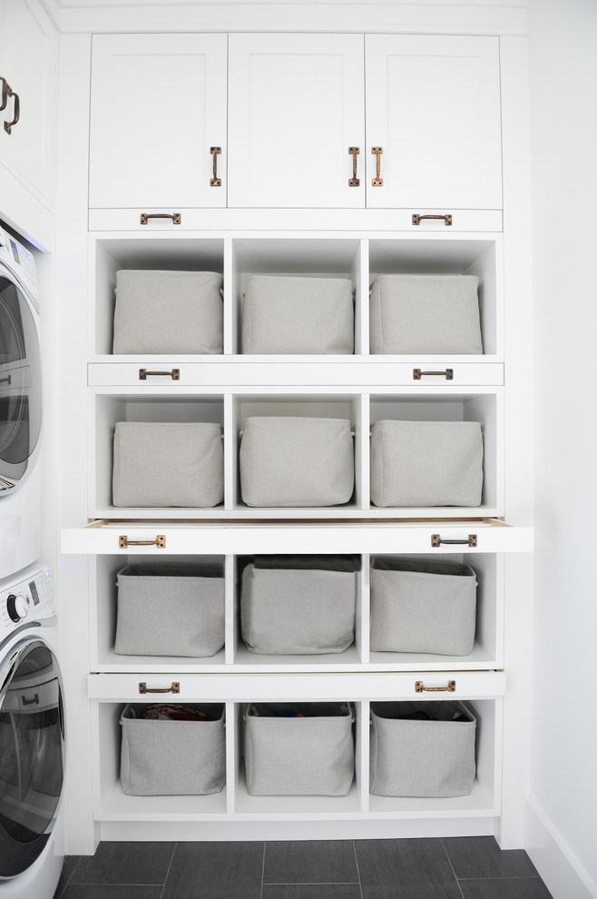 This is an example of a large country l-shaped dedicated laundry room in Vancouver with white cabinets, white walls, a stacked washer and dryer, open cabinets, ceramic floors and grey floor.
