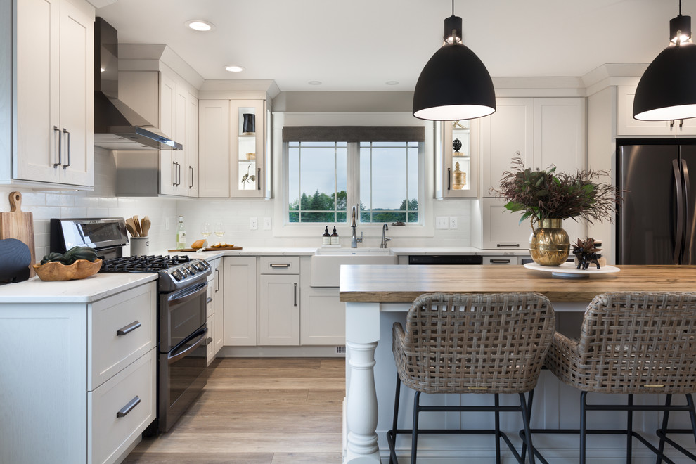 Inspiration for a transitional l-shaped kitchen in Other with a farmhouse sink, shaker cabinets, white cabinets, white splashback, stainless steel appliances, medium hardwood floors, with island, brown floor and white benchtop.