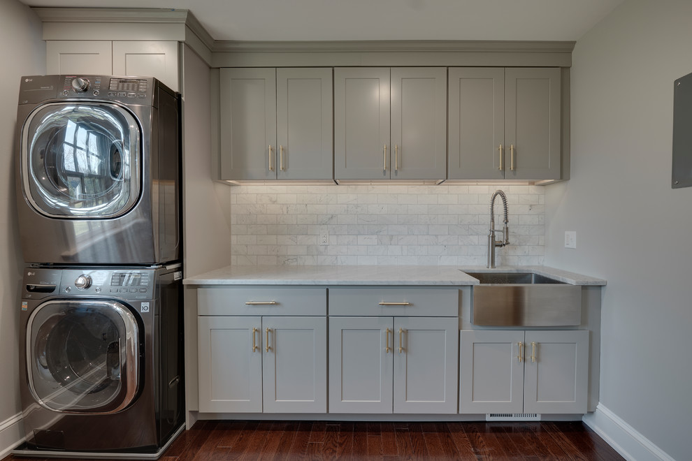 Design ideas for a mid-sized transitional single-wall dedicated laundry room in Philadelphia with a farmhouse sink, recessed-panel cabinets, grey cabinets, marble benchtops, grey walls, medium hardwood floors, a stacked washer and dryer, brown floor and white benchtop.