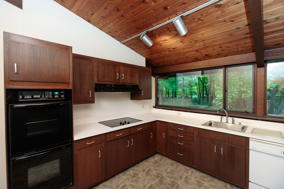 Photo of a mid-sized midcentury galley eat-in kitchen in Boston with a drop-in sink, flat-panel cabinets, dark wood cabinets, solid surface benchtops, white splashback, black appliances, ceramic floors, beige floor and white benchtop.