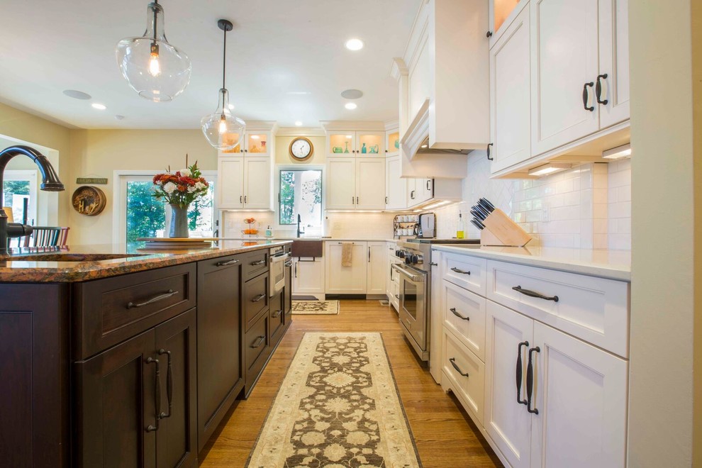 Example of a large transitional l-shaped light wood floor and brown floor enclosed kitchen design with a farmhouse sink, shaker cabinets, white cabinets, solid surface countertops, white backsplash, porcelain backsplash, stainless steel appliances and an island