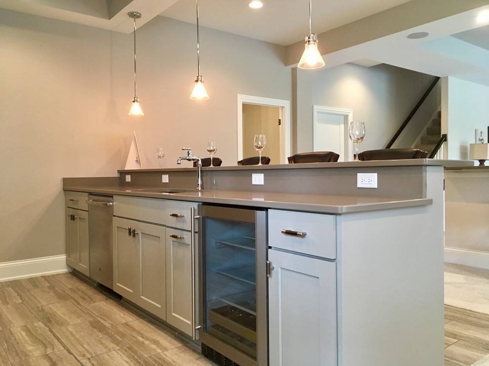 Photo of a large transitional galley wet bar in Milwaukee with an undermount sink, recessed-panel cabinets, grey cabinets, quartz benchtops, grey splashback, ceramic floors, grey floor and grey benchtop.