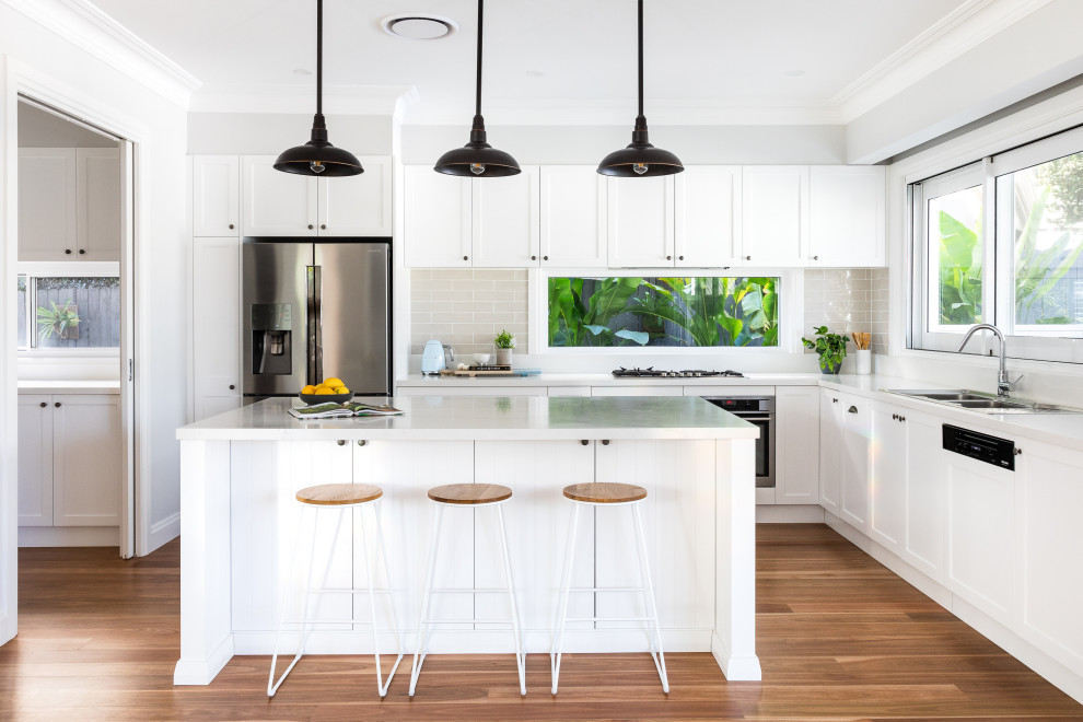 This is an example of a mid-sized beach style l-shaped kitchen with a double-bowl sink, shaker cabinets, white cabinets, quartz benchtops, light hardwood floors, with island, white benchtop, grey splashback, subway tile splashback, stainless steel appliances and beige floor.