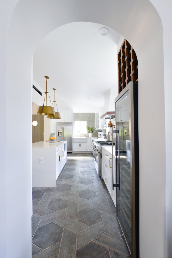 Example of a mid-sized minimalist l-shaped porcelain tile and gray floor kitchen design in Miami with a farmhouse sink, shaker cabinets, white cabinets, quartz countertops, gray backsplash, brick backsplash, stainless steel appliances and an island