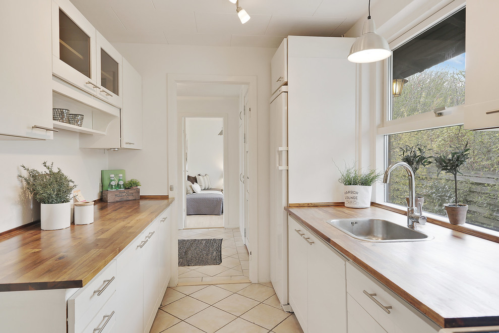 This is an example of a scandinavian galley kitchen in Copenhagen with a drop-in sink, flat-panel cabinets, white cabinets, wood benchtops, white splashback, window splashback, white appliances and beige floor.