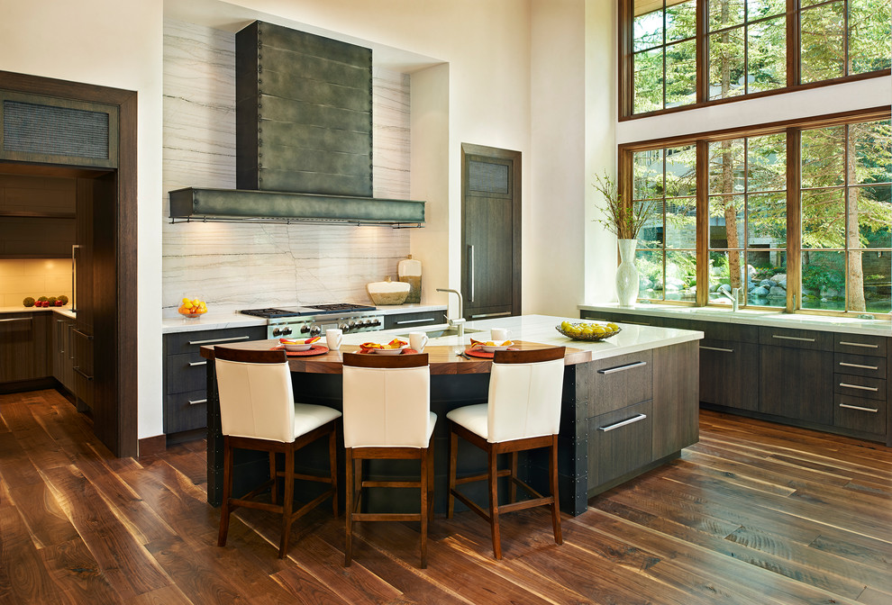 This is an example of a transitional l-shaped eat-in kitchen in Denver with flat-panel cabinets, dark wood cabinets, medium hardwood floors, with island, an undermount sink, solid surface benchtops, beige splashback, porcelain splashback and stainless steel appliances.