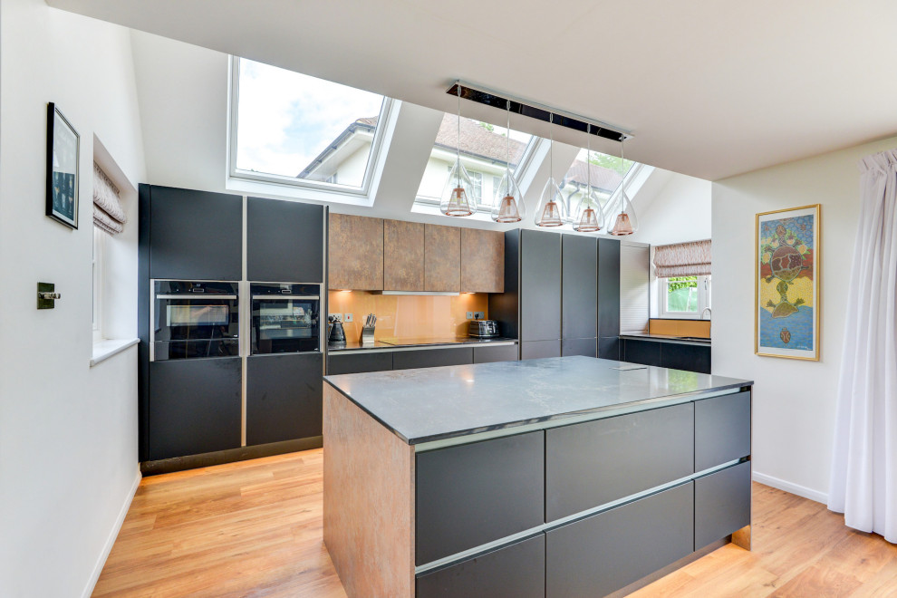 This is an example of a large modern grey and black l-shaped open plan kitchen in Sussex with an integrated sink, flat-panel cabinets, black cabinets, quartz worktops, brown splashback, glass sheet splashback, black appliances, laminate floors, an island, brown floors, black worktops, a vaulted ceiling and feature lighting.