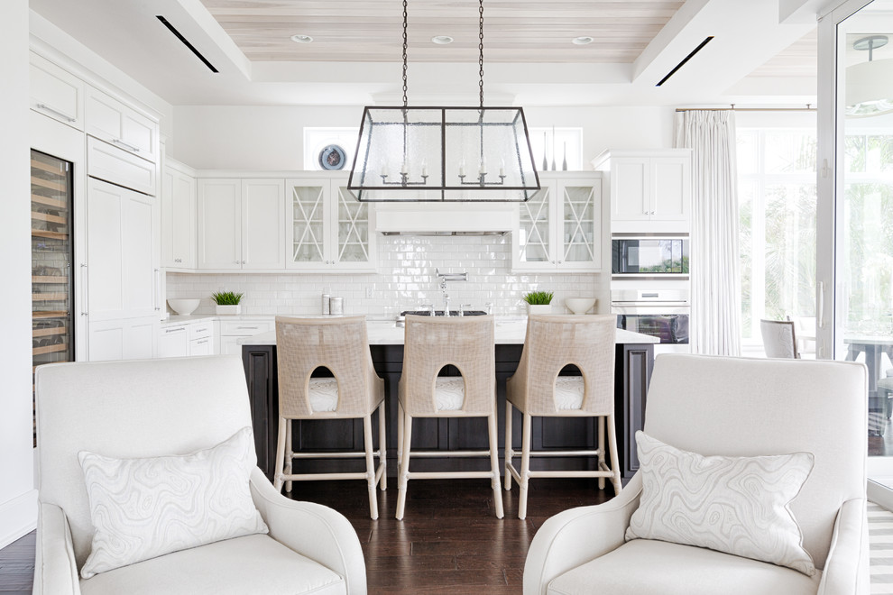 Beach style l-shaped open plan kitchen in Miami with glass-front cabinets, white cabinets, white splashback, subway tile splashback, panelled appliances, dark hardwood floors, with island and brown floor.