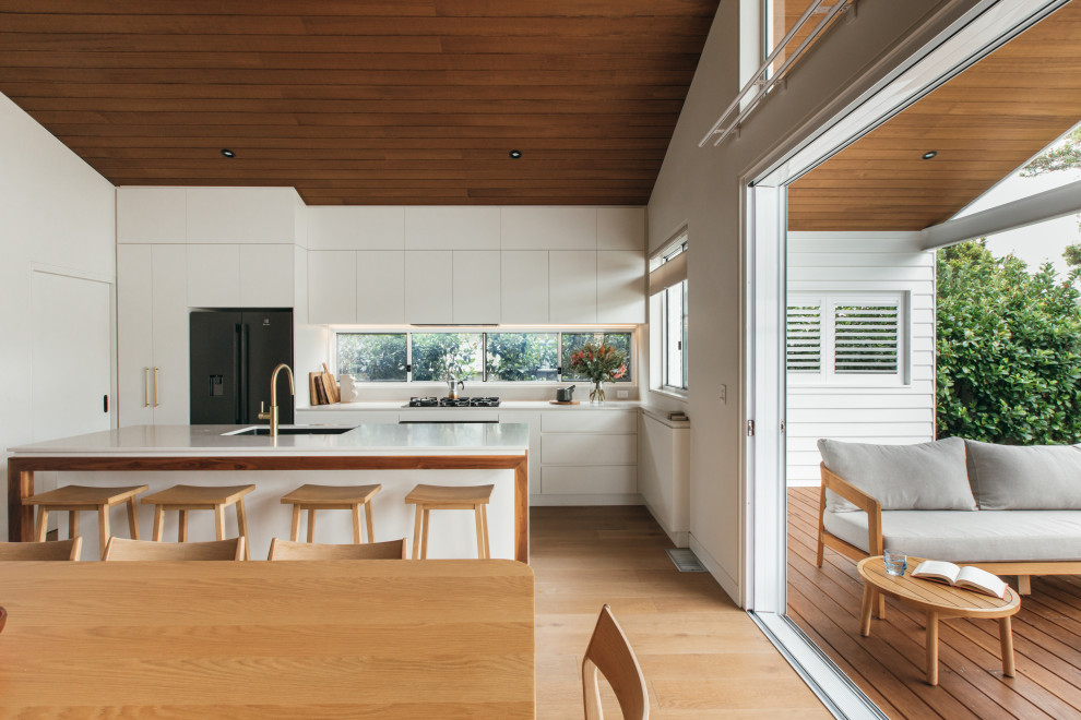 Inspiration for a midcentury l-shaped eat-in kitchen in Central Coast with an undermount sink, flat-panel cabinets, white cabinets, window splashback, stainless steel appliances, medium hardwood floors, with island, brown floor, white benchtop, vaulted and wood.
