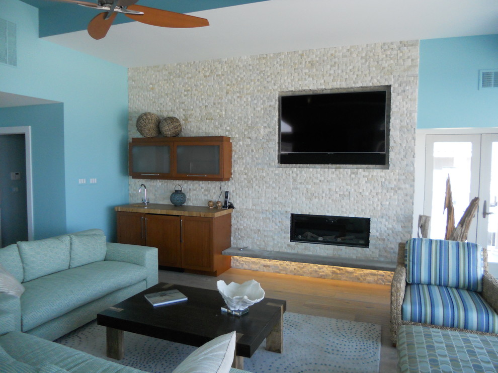 This is an example of a large beach style open concept living room in New York with a home bar, blue walls, light hardwood floors, a ribbon fireplace, a stone fireplace surround and grey floor.