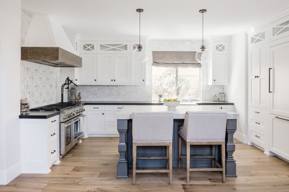 Example of a transitional u-shaped light wood floor and beige floor kitchen design in Orange County with a farmhouse sink, shaker cabinets, white cabinets, white backsplash, paneled appliances, an island and black countertops