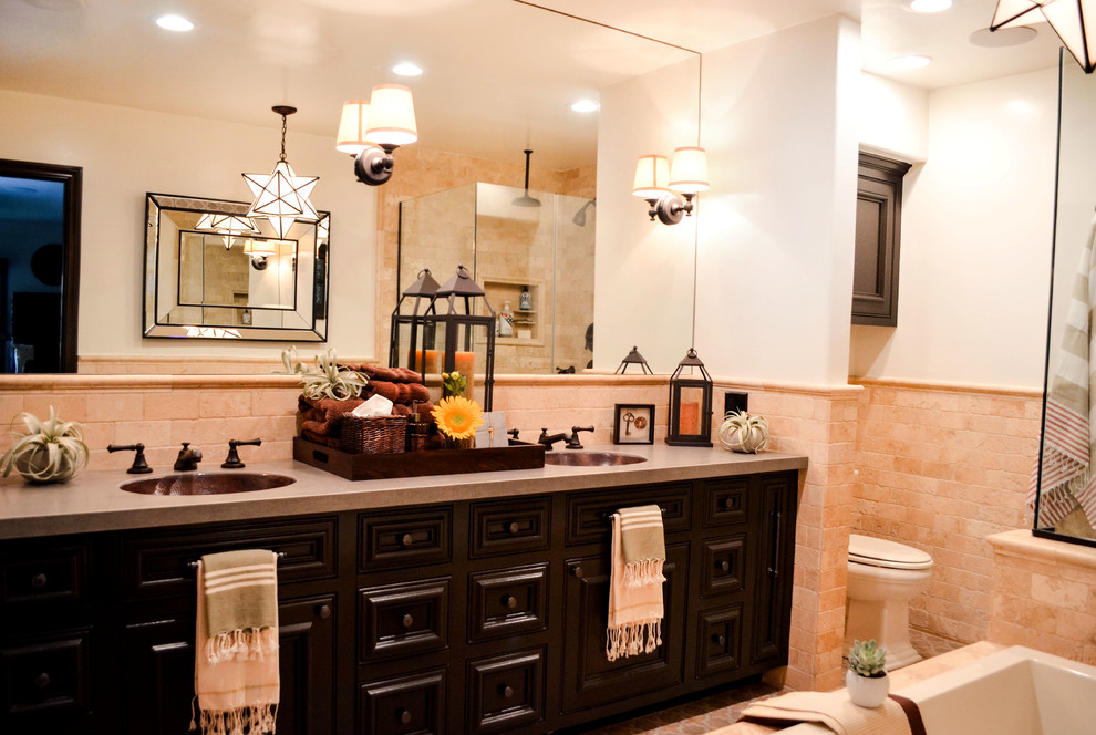 This is an example of a mid-sized mediterranean master bathroom in Los Angeles with beaded inset cabinets, brown cabinets, a drop-in tub, a corner shower, a one-piece toilet, beige tile, travertine, white walls, porcelain floors, an undermount sink, solid surface benchtops, multi-coloured floor, a hinged shower door and grey benchtops.
