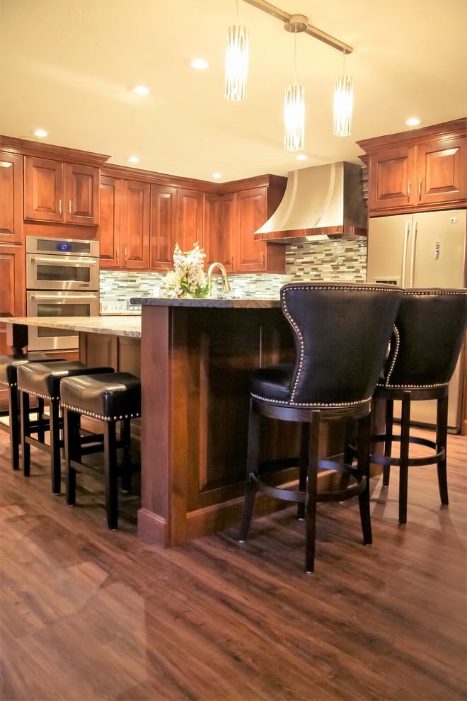 Large transitional l-shaped medium tone wood floor eat-in kitchen photo with an undermount sink, raised-panel cabinets, medium tone wood cabinets, granite countertops, glass tile backsplash, stainless steel appliances and an island
