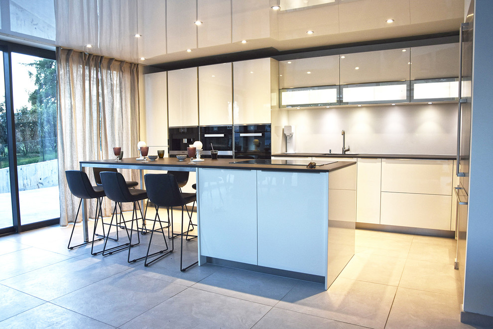 This is an example of a large modern l-shaped open plan kitchen in Lyon with an undermount sink, white cabinets, white splashback, with island, grey floor, beaded inset cabinets, limestone benchtops, glass sheet splashback, black appliances and ceramic floors.