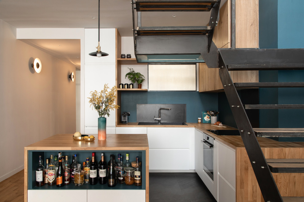 This is an example of an industrial l-shaped kitchen in Paris with a drop-in sink, flat-panel cabinets, white cabinets, wood benchtops, stainless steel appliances, with island, black floor and brown benchtop.