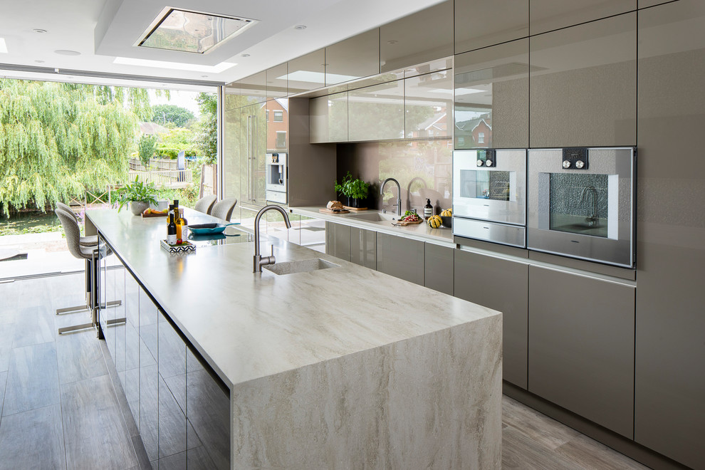 Photo of a mid-sized contemporary galley kitchen in Other with flat-panel cabinets, grey cabinets, solid surface benchtops, glass sheet splashback, stainless steel appliances, porcelain floors, with island, multi-coloured floor, beige benchtop, an integrated sink and beige splashback.