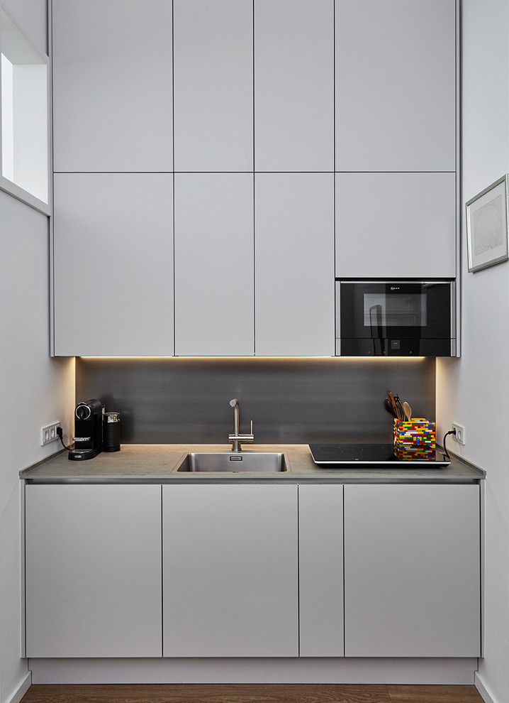 Photo of a small contemporary single-wall eat-in kitchen in Dusseldorf with a single-bowl sink, flat-panel cabinets, grey cabinets, solid surface benchtops, metallic splashback, metal splashback, black appliances, medium hardwood floors, brown floor and no island.