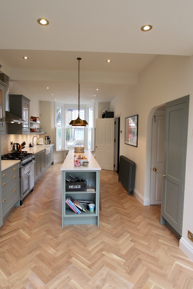 Mid-sized contemporary single-wall eat-in kitchen in London with a farmhouse sink, shaker cabinets, green cabinets, solid surface benchtops, white splashback, stainless steel appliances, light hardwood floors, with island, brown floor and white benchtop.