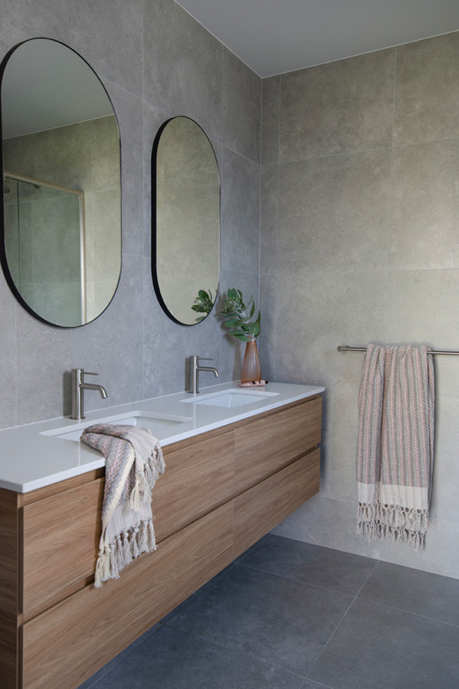 Пример оригинального дизайна: ванная комната среднего размера в стиле модернизм с фасадами островного типа, светлыми деревянными фасадами, угловым душем, унитазом-моноблоком, серой плиткой, керамогранитной плиткой, серыми стенами, полом из керамогранита, душевой кабиной, накладной раковиной, столешницей из искусственного кварца, серым полом, душем с распашными дверями, белой столешницей, тумбой под две раковины и подвесной тумбой