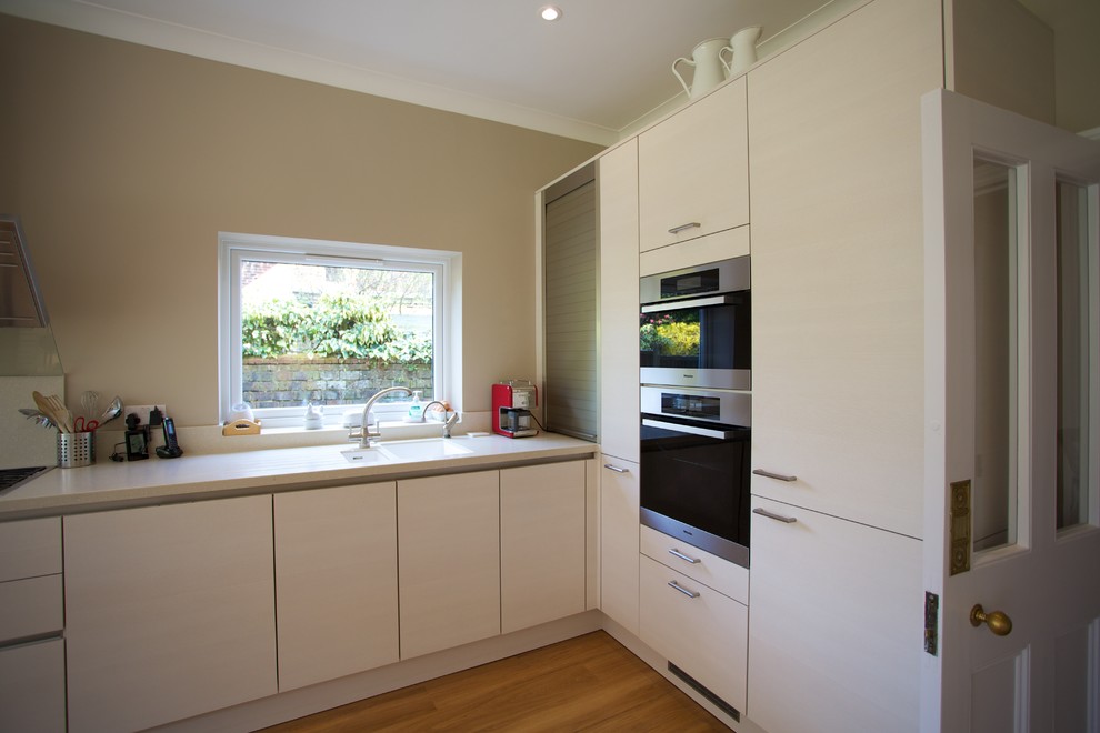 Inspiration for a mid-sized contemporary u-shaped eat-in kitchen in Hampshire with an integrated sink, flat-panel cabinets, white cabinets, solid surface benchtops, white splashback, stainless steel appliances, medium hardwood floors and a peninsula.