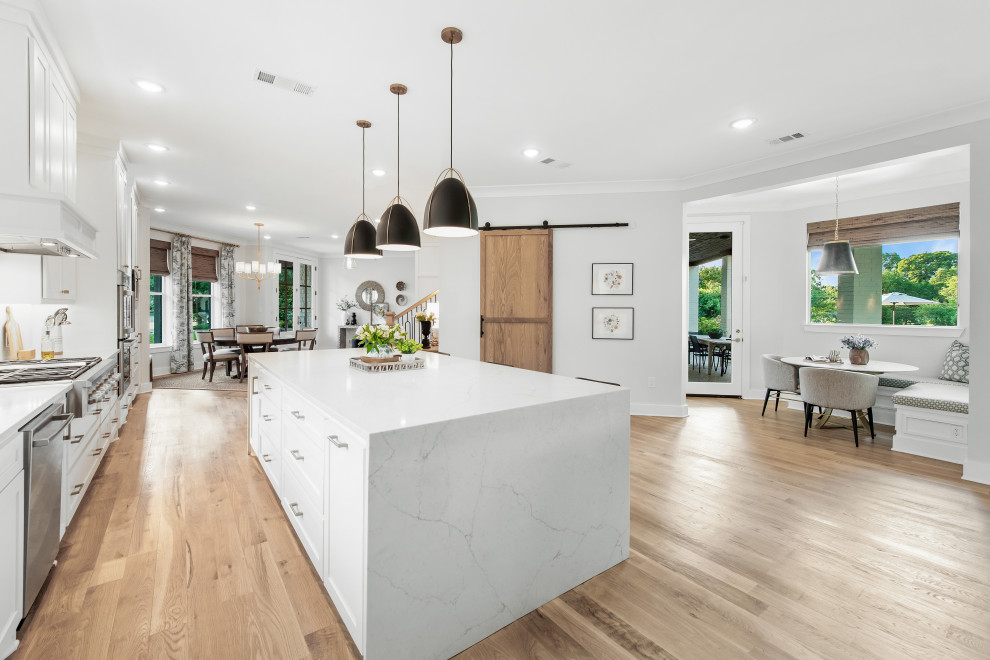 Example of a large transitional single-wall light wood floor open concept kitchen design with a farmhouse sink, shaker cabinets, white cabinets, quartz countertops, white backsplash, ceramic backsplash, stainless steel appliances, an island and white countertops