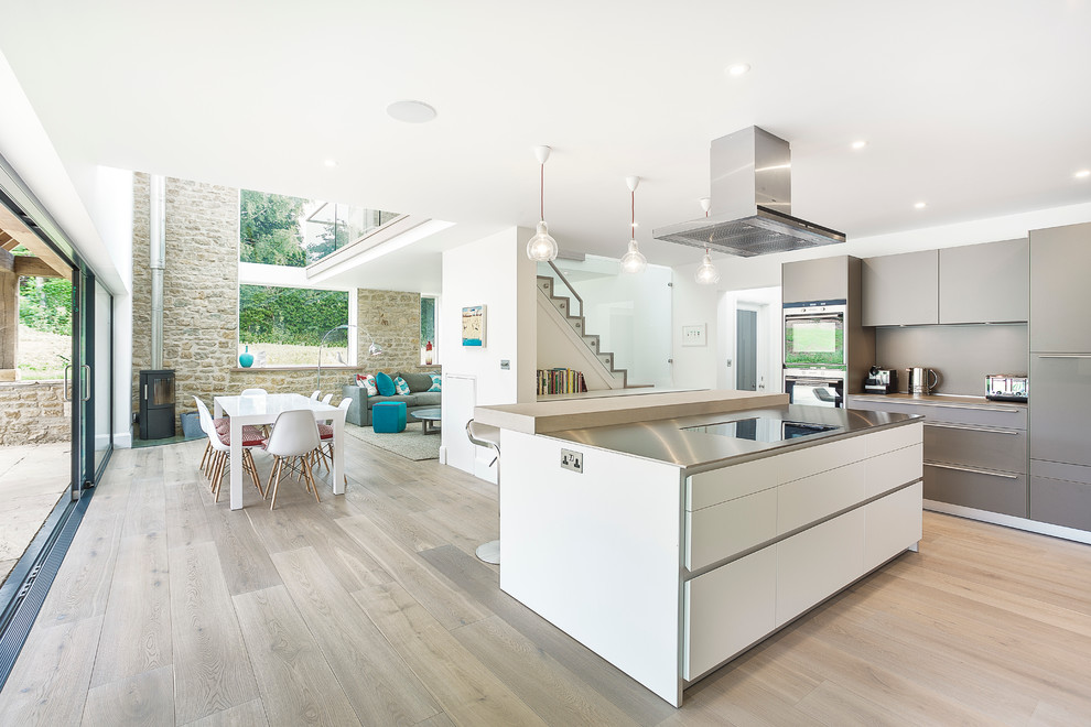Inspiration for a mid-sized contemporary l-shaped open plan kitchen in Hampshire with flat-panel cabinets, grey cabinets, stainless steel benchtops, grey splashback, stainless steel appliances, light hardwood floors and with island.