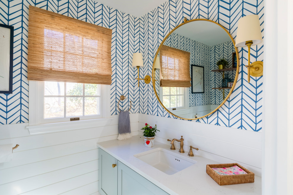 Photo of a mid-sized beach style powder room in New York with shaker cabinets, blue cabinets, a two-piece toilet, blue walls, marble floors, engineered quartz benchtops, white floor, white benchtops, a floating vanity and wallpaper.