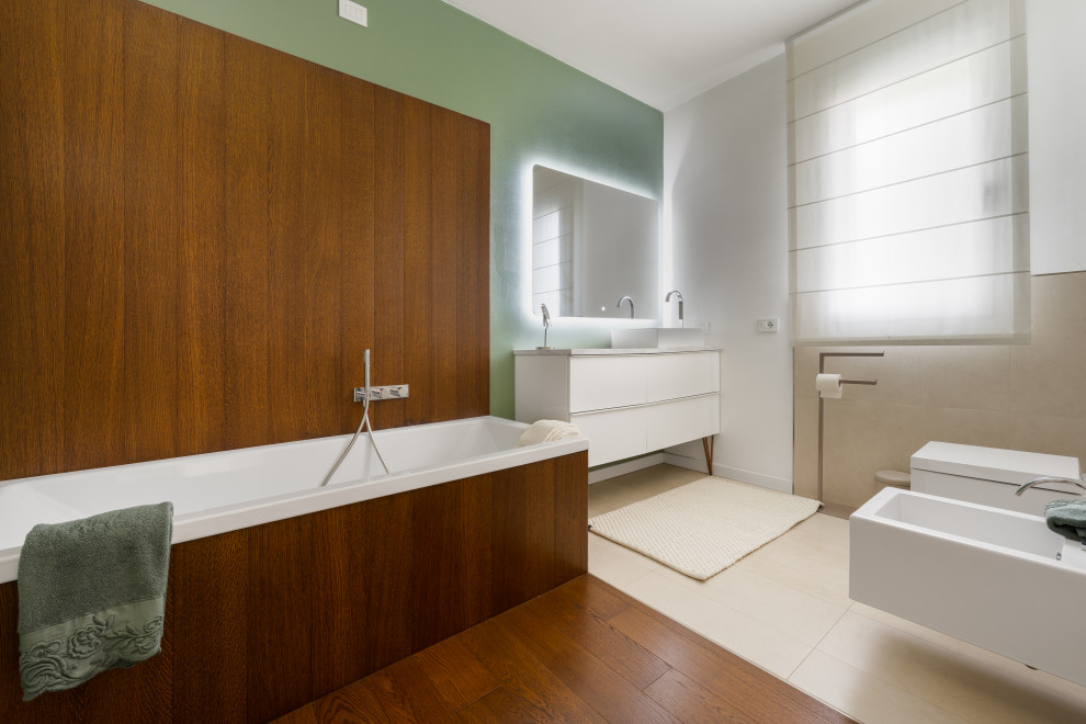 This is an example of a modern master bathroom in Venice with white cabinets, a drop-in tub, a wall-mount toilet, beige tile, green walls, porcelain floors, a vessel sink, marble benchtops, beige floor, grey benchtops, an enclosed toilet, a freestanding vanity and wood walls.