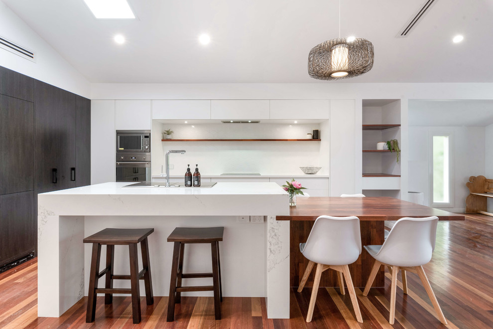 Photo of a contemporary l-shaped kitchen in Brisbane with flat-panel cabinets, dark wood cabinets, white splashback, stainless steel appliances, medium hardwood floors, with island, brown floor and white benchtop.