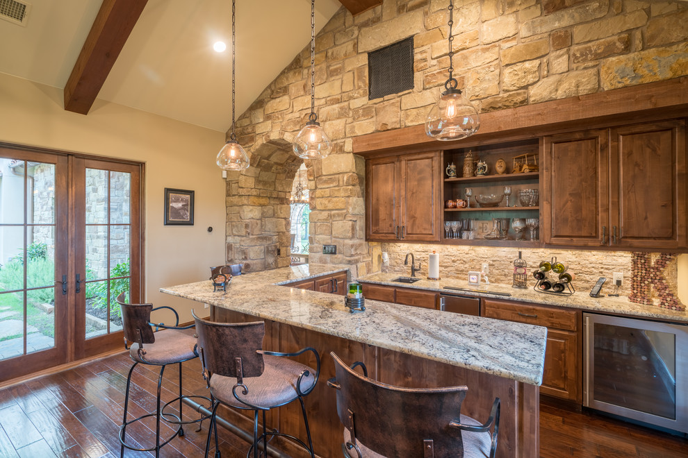 This is an example of a mid-sized country u-shaped seated home bar in Austin with an undermount sink, raised-panel cabinets, medium wood cabinets, beige splashback, medium hardwood floors, granite benchtops, stone tile splashback and brown floor.