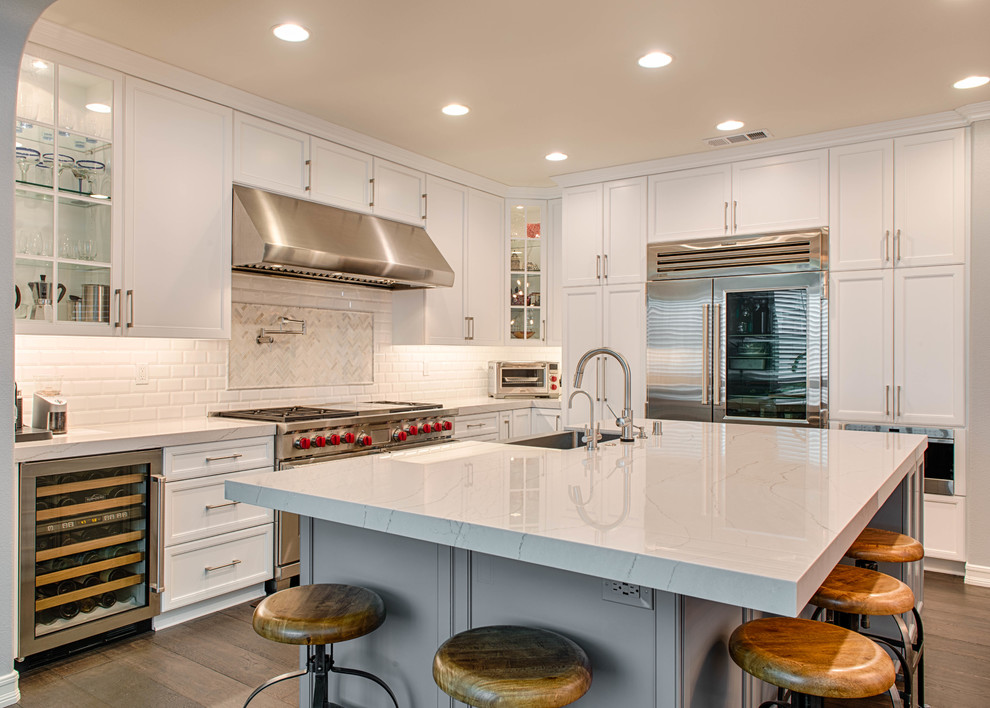 Design ideas for a transitional l-shaped kitchen in Los Angeles with a farmhouse sink, shaker cabinets, white cabinets, white splashback, subway tile splashback, stainless steel appliances, dark hardwood floors and with island.