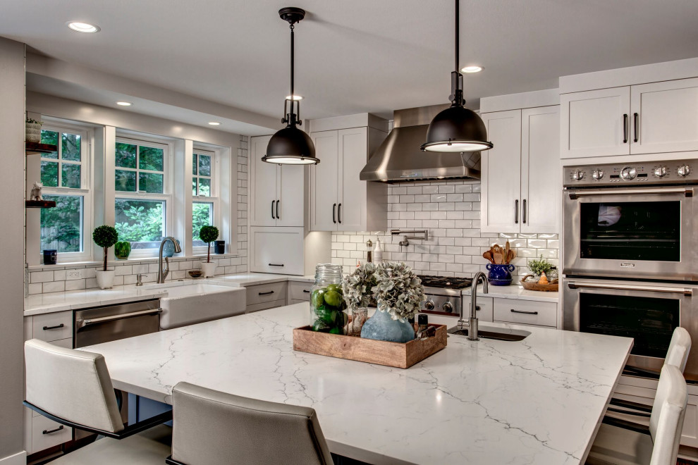 Design ideas for a transitional l-shaped kitchen in Seattle with a farmhouse sink, shaker cabinets, white cabinets, white splashback, subway tile splashback, stainless steel appliances, with island and white benchtop.