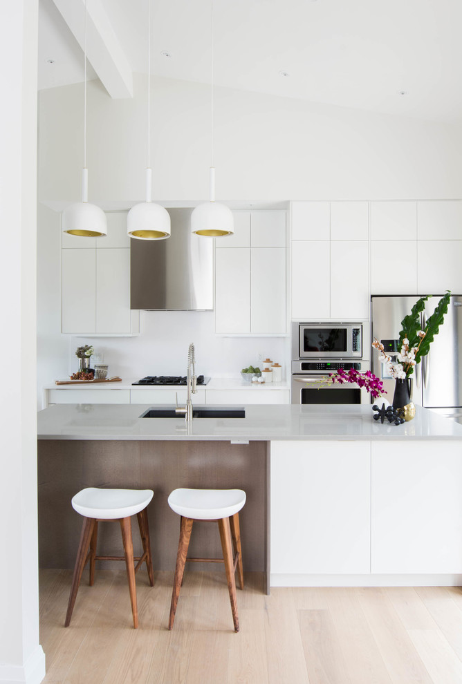 This is an example of a mid-sized scandinavian galley open plan kitchen in Toronto with an undermount sink, flat-panel cabinets, white cabinets, quartzite benchtops, white splashback, stainless steel appliances, light hardwood floors and with island.