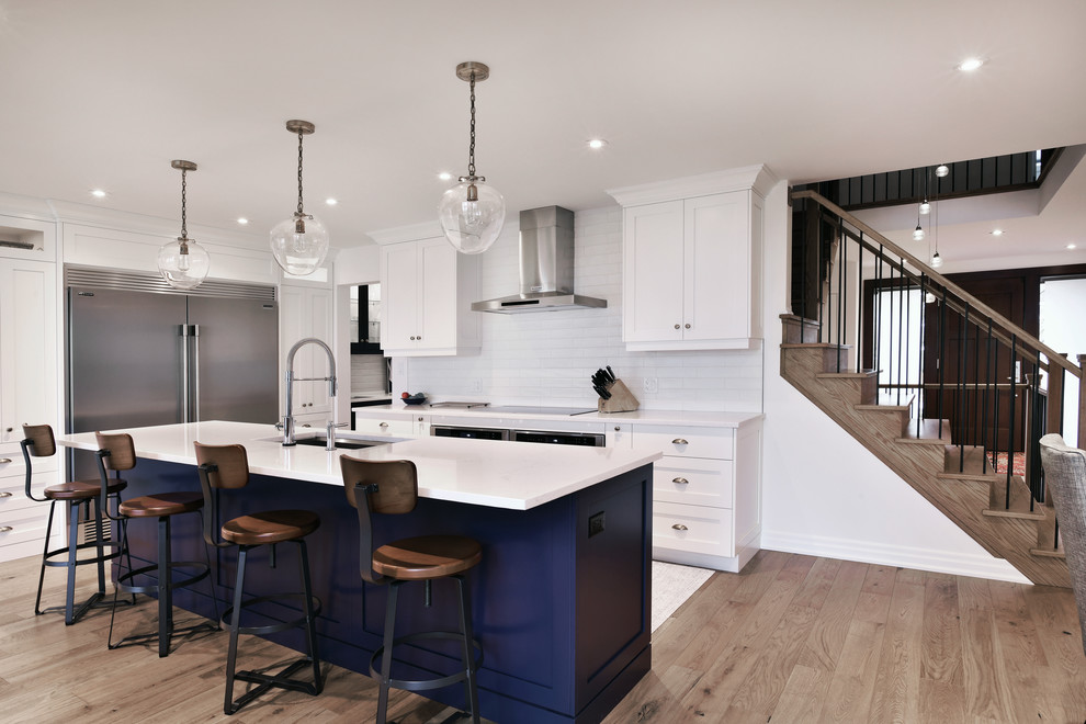 Inspiration for a large beach style galley kitchen in Toronto with an undermount sink, recessed-panel cabinets, white cabinets, quartz benchtops, white splashback, cement tile splashback, stainless steel appliances, light hardwood floors, with island, brown floor and white benchtop.
