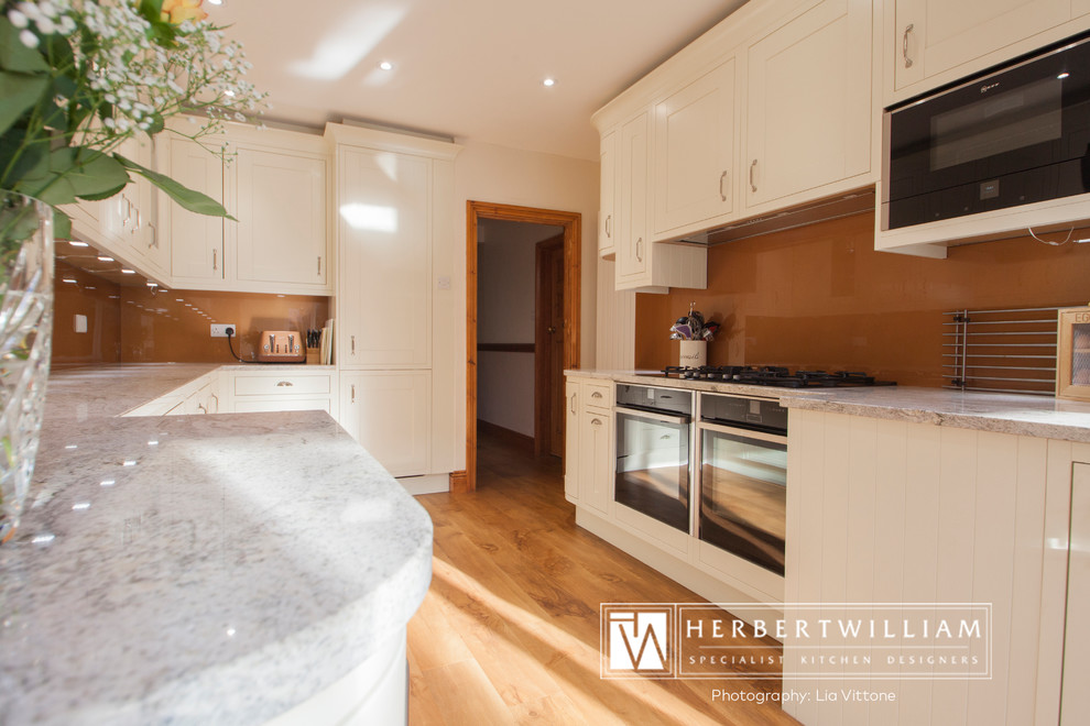 This is an example of a large traditional l-shaped eat-in kitchen in Hampshire with shaker cabinets, white cabinets, glass benchtops, brown splashback, glass sheet splashback, stainless steel appliances, medium hardwood floors, a peninsula and brown floor.